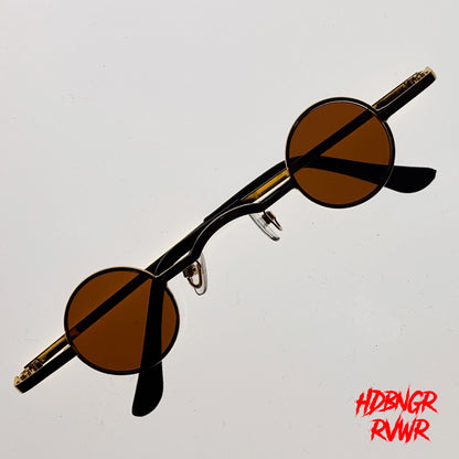 Round-frame sunglasses with amber-brown lenses and black temples, photographed on a plain light background. A red stylized text watermark reading 'HDBNGR RNWR' appears in the bottom-right corner.