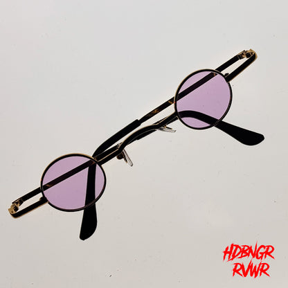Round metal-framed sunglasses with light purple tinted lenses and black temple arms, photographed on a plain light background with a red stylized text watermark in the bottom right corner.