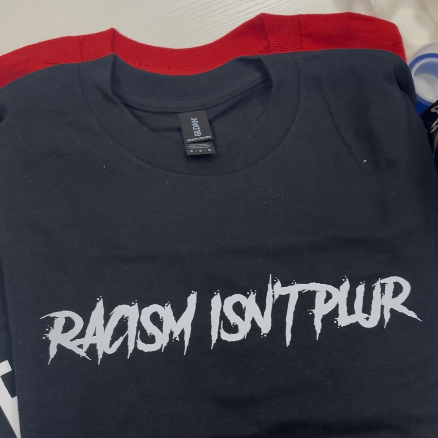 A folded black t-shirt with a red collar band, featuring the text 'RACISM ISN'T PLUR' printed in a stylized white font across the chest.