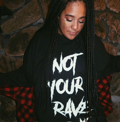 A woman with long black box braids wears a black oversized T-shirt with distressed white text reading 'NOT YOUR RAVE' and a red-and-black plaid shirt draped off her shoulders, posed against a rustic stone wall.